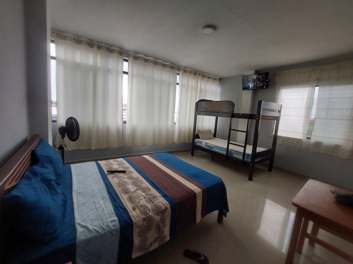 A room is featured with a spacious bed dressed in blue and brown linens. A bunk bed sits against the wall, and a small table is positioned near a window. Natural light filters in through multiple large windows adorned with light curtains.