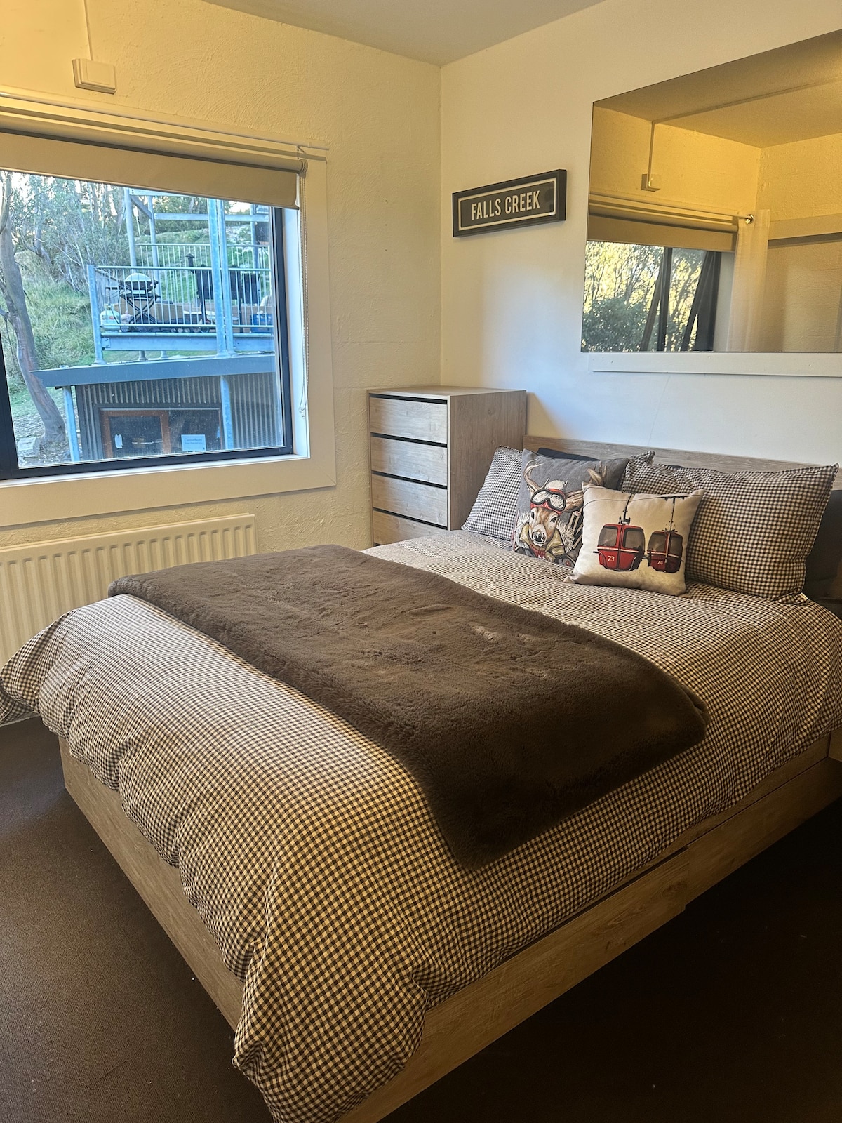 A cozy sleeping area features a spacious bed covered with a checkered duvet and a soft brown throw. Decorative pillows enhance the design, while a dresser stands nearby. Natural light filters in through a window that overlooks the surrounding greenery.