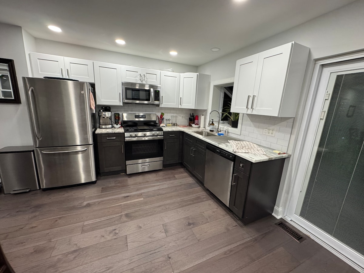 A modern kitchen is seen with sleek white cabinetry and stainless steel appliances, including a refrigerator, microwave, and oven. The countertop is finished with a light-colored stone, providing a bright workspace. A sliding glass door offers access to an outdoor area.