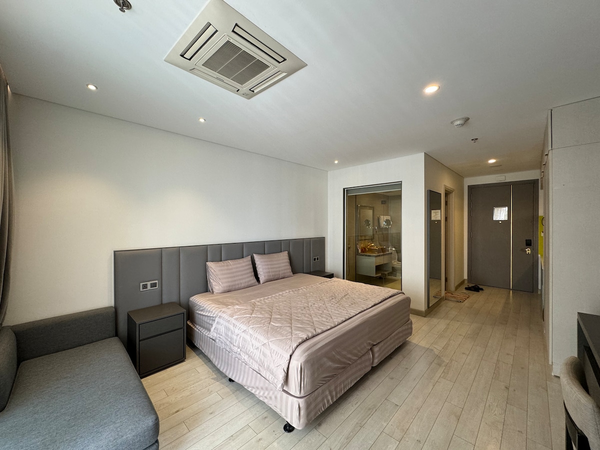 A spacious studio layout features a large, comfortable bed with a soft, muted coverlet. A sleek gray sofa sits beside the bed, and glass doors provide a view into a partially visible bathroom area. Natural light fills the room through recessed ceiling lights.
