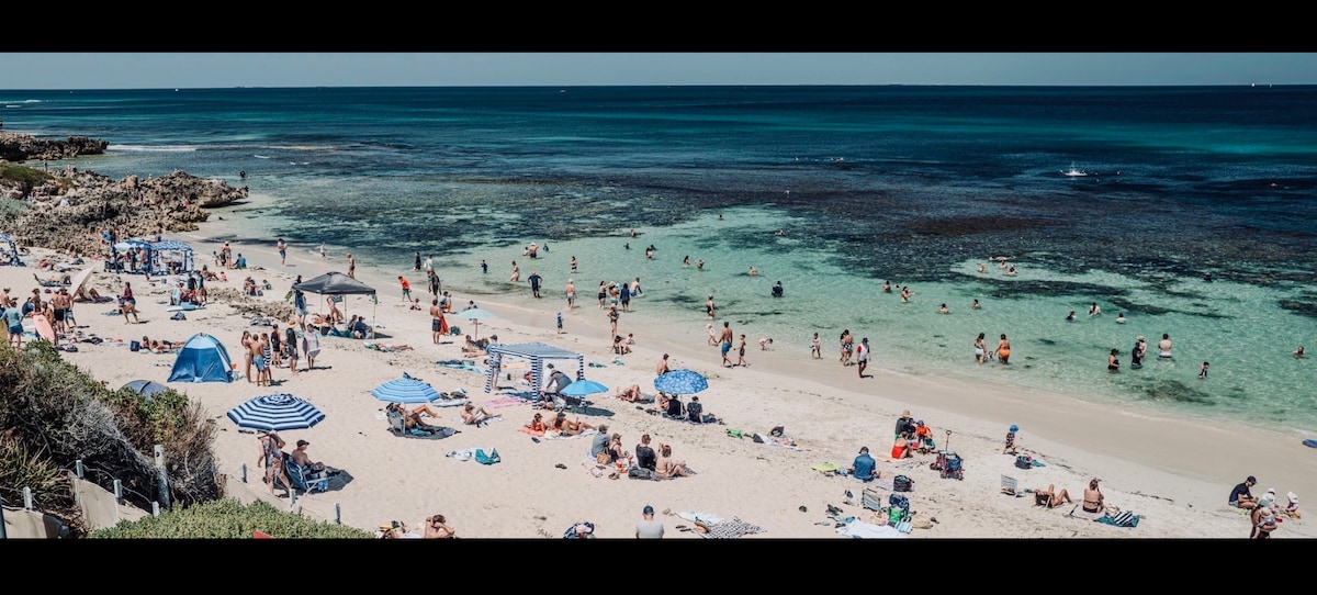 A vibrant beach scene is depicted, featuring a mix of sunbathers and swimmers enjoying the clear turquoise waters. Umbrellas and towels are scattered across the sandy shore, with rocky formations visible on the left. The expansive ocean and a clear sky create a refreshing coastal atmosphere.