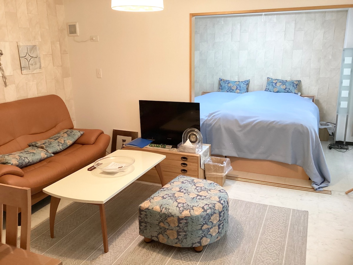 A compact living area features a brown leather sofa adorned with floral cushions. A wooden table sits in front, accompanied by a light blue blanket draped over a spacious bed in the background. A TV stands on a low cabinet, with additional items organized nearby.