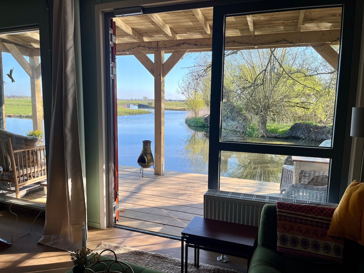 A view from inside showcases a spacious patio with wooden beams, overlooking a serene body of water and lush greenery. Large glass doors allow natural light to fill the space, creating a harmonious connection between indoor comfort and the calming outdoor environment.