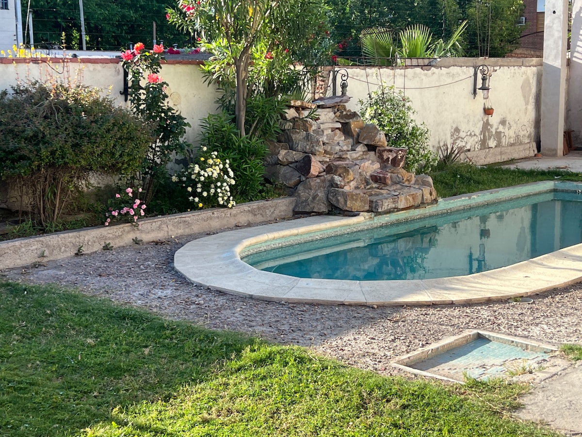 A serene outdoor area features a well-maintained pool surrounded by a landscaped garden. A natural stone fountain adds a decorative touch beside lush greenery and blooming flowers. The pool's clear water reflects the surrounding plants and sunlight, creating a calm atmosphere.