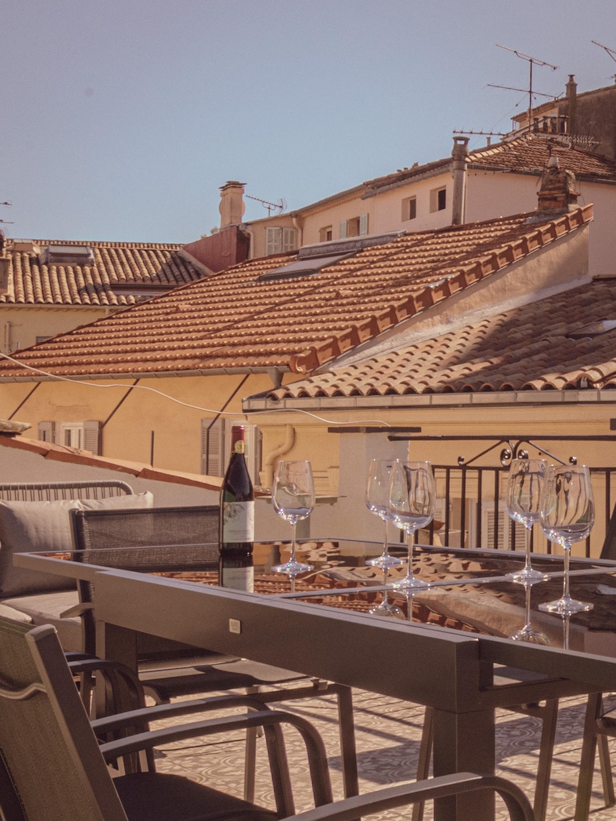 A private rooftop terrace is featured, showcasing a dining table surrounded by six chairs. Five wine glasses are placed on the table, along with a bottle of sparkling wine. Traditional rooftops with terracotta tiles can be seen in the background beneath a clear blue sky.