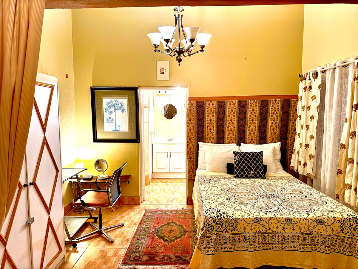 A cozy sleeping area features a spacious bed with patterned bedding, accented by decorative pillows. A desk and chair are positioned beside the bed. Soft lighting is provided by a chandelier overhead, while patterned curtains frame the windows and warm tones enhance the inviting atmosphere.