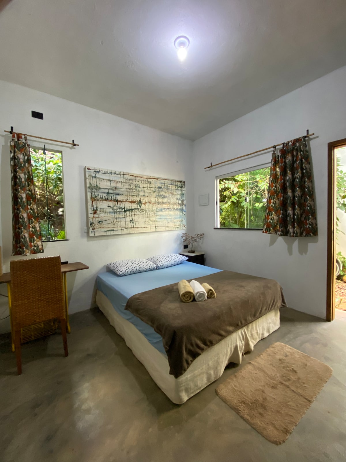 A cozy suite features a spacious bed with a brown blanket and white pillows. A wooden work desk is positioned nearby. Large windows invite natural light, showcasing lush greenery outside. Floral curtains frame the windows, adding a touch of color to the neutral walls.