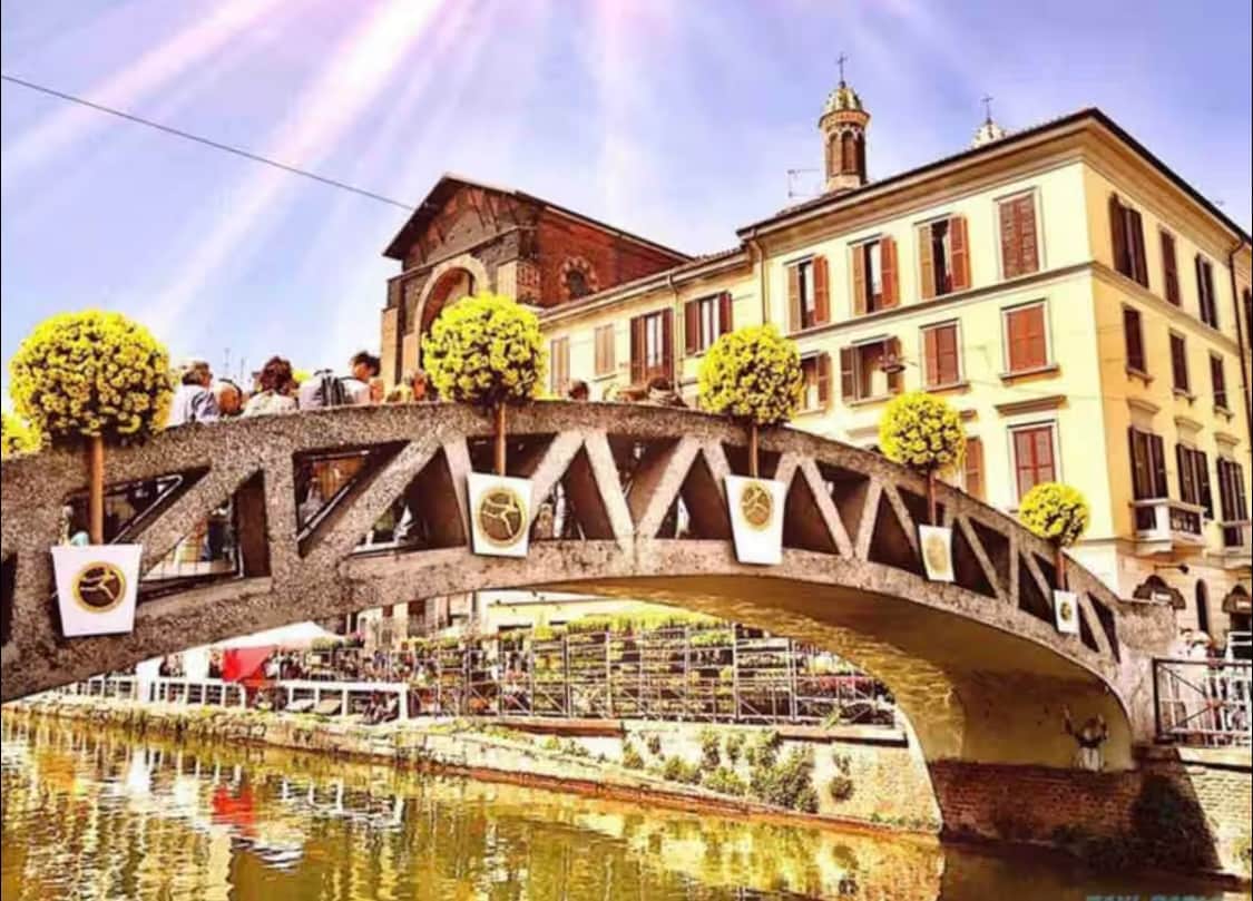 A charming bridge adorned with round, trimmed trees spans a canal, connecting lively spaces on either side. Historic buildings featuring a mix of architectural styles are visible in the background, with outdoor seating areas inviting relaxation along the water's edge.