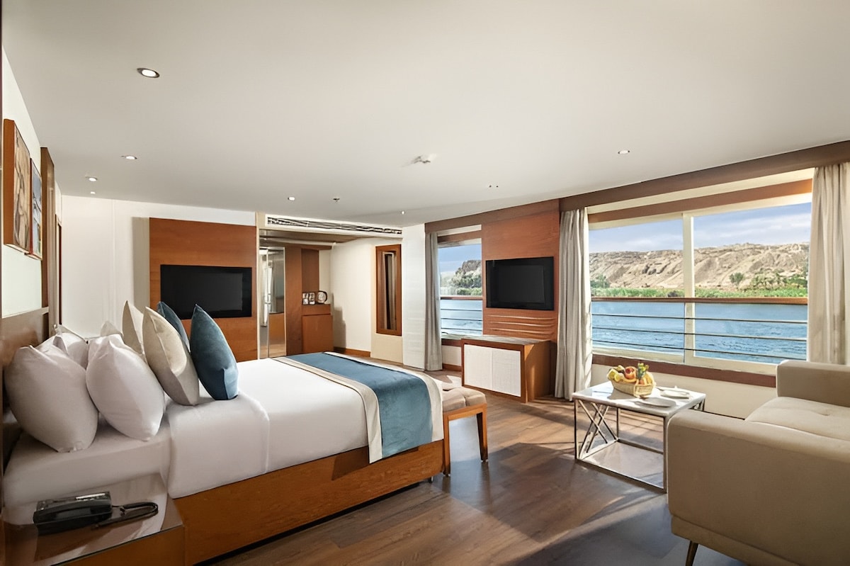 A spacious cabin is presented with large windows offering views of the Nile. The room features a comfortable bed with neatly arranged pillows and a cozy seating area, complete with a modern coffee table. Two flat-screen televisions are mounted on the wall.