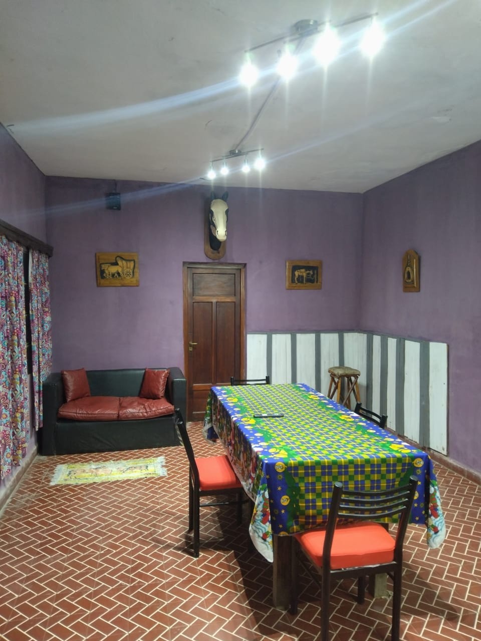 A relaxed dining area features a colorful tablecloth covering a long table surrounded by five chairs. A dark couch is positioned along one wall, complemented by patterned curtains. The space is illuminated by overhead lights, enhancing the cozy atmosphere.