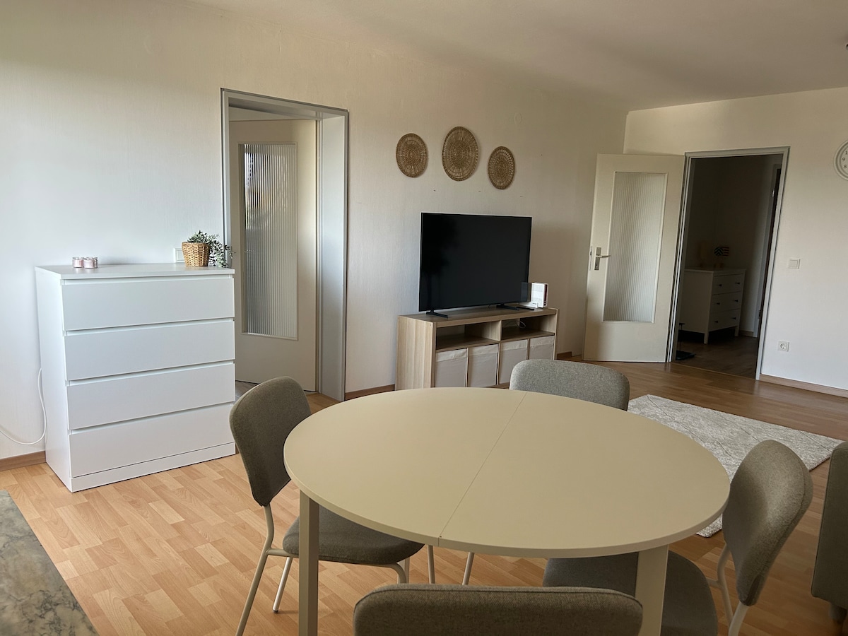 A bright living area features a circular dining table surrounded by four chairs. A television is positioned on a low wooden stand, and a dresser is visible in the background. Light wood flooring complements the neutral wall tones, enhancing the open atmosphere.