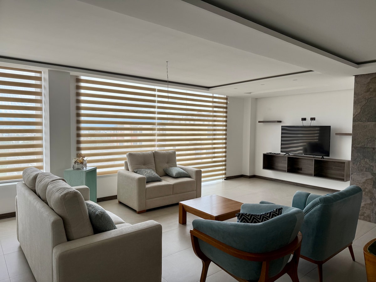 A spacious living area features large windows with horizontal blinds allowing natural light to filter in. Two light-toned sofas and two teal chairs are arranged around a wooden coffee table. A flat-screen television is mounted on the wall, complemented by simple shelving beneath.
