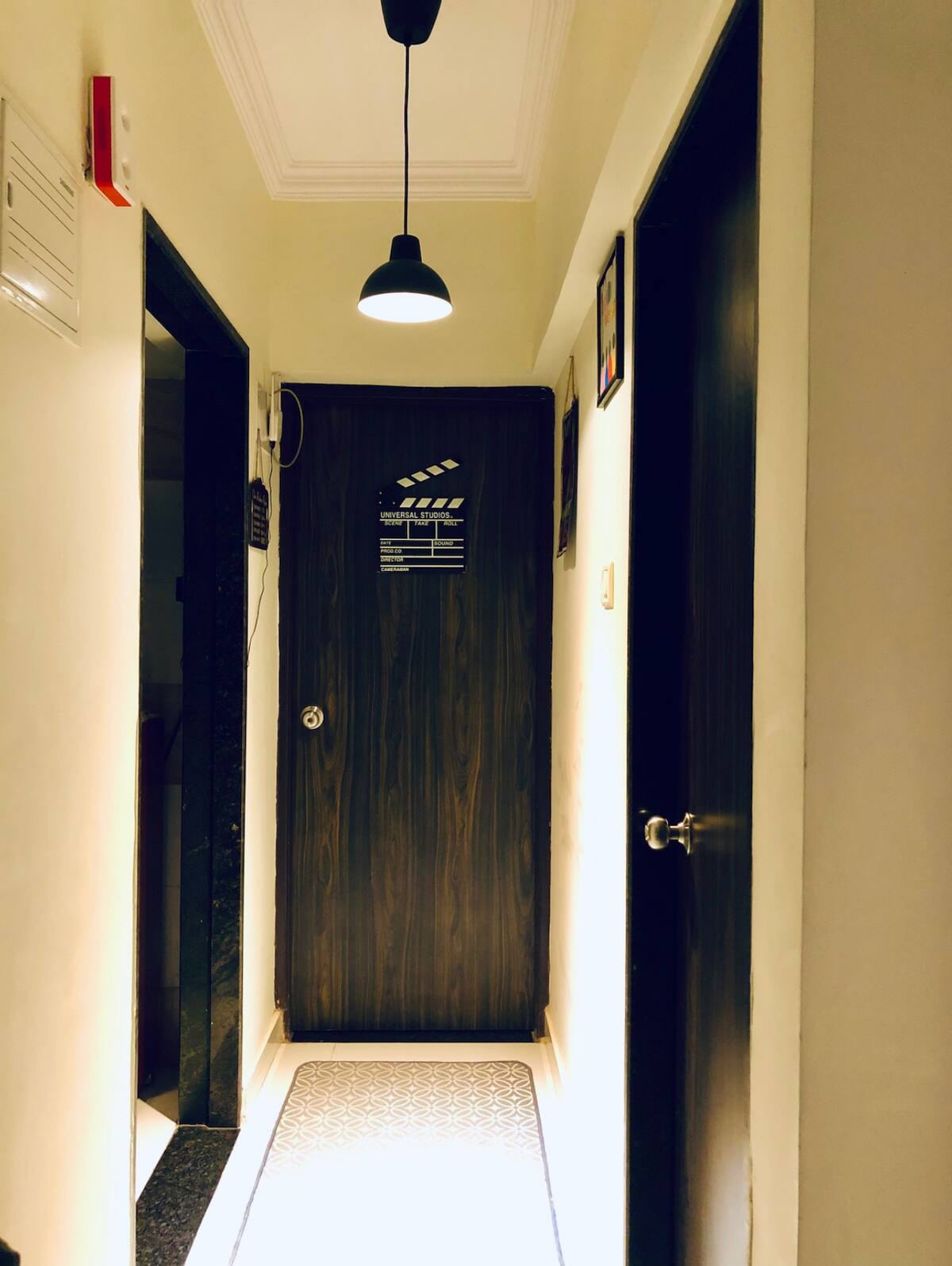A well-lit hallway is depicted, featuring light-colored walls and a dark wood door at the end. A pendant light hangs from the ceiling, illuminating the space. Decorative frames are visible on the wall, and a patterned runner mat extends along the floor.