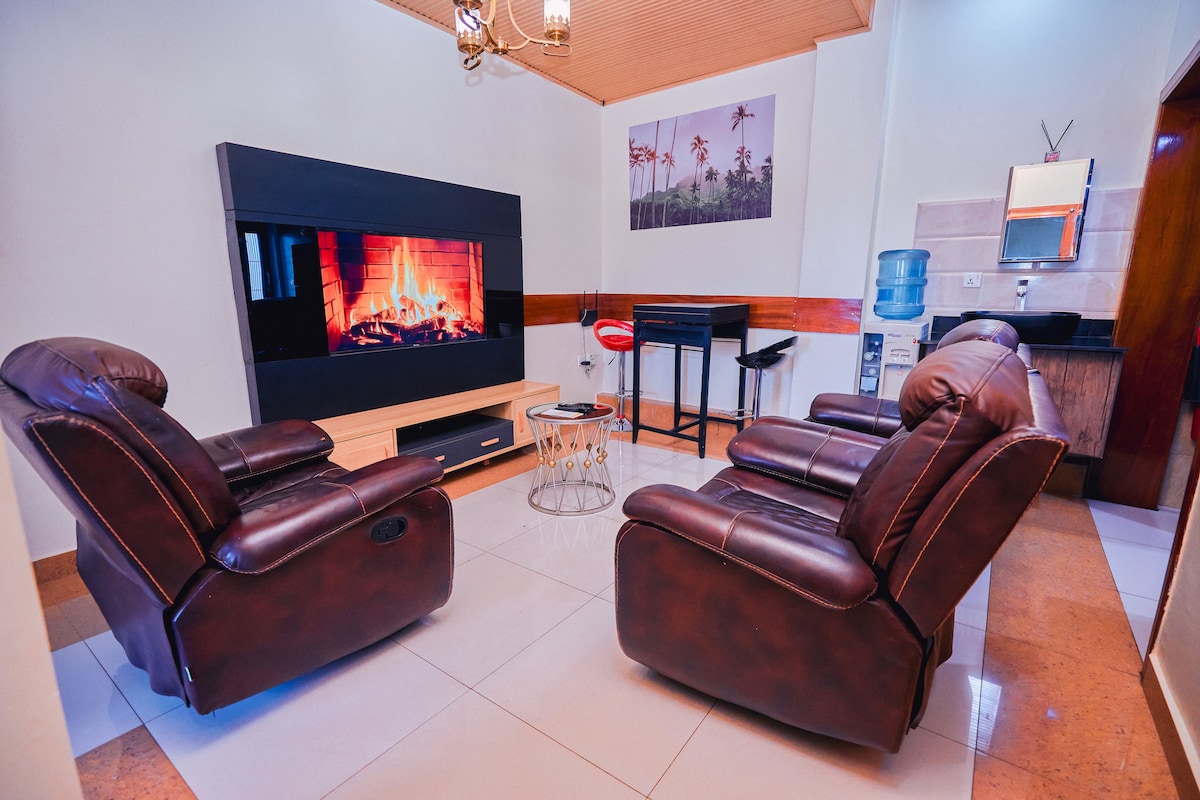 A comfortable seating area is presented, featuring two recliners in a rich brown leather. A modern television is mounted on the wall, above a sleek entertainment unit. A small glass-topped table sits between the chairs, and a water cooler can be seen in the corner.