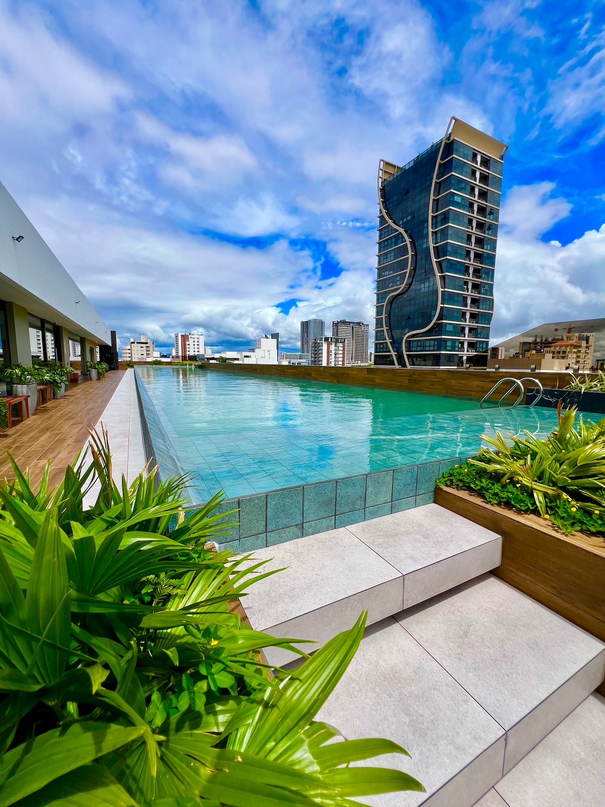 A sleek rooftop pool is framed by modern architecture, with sparkling water reflecting the blue sky. Lush greenery borders the pool, while steps lead down from the deck. The skyline of Santa Cruz is visible in the background, enhancing the urban atmosphere.