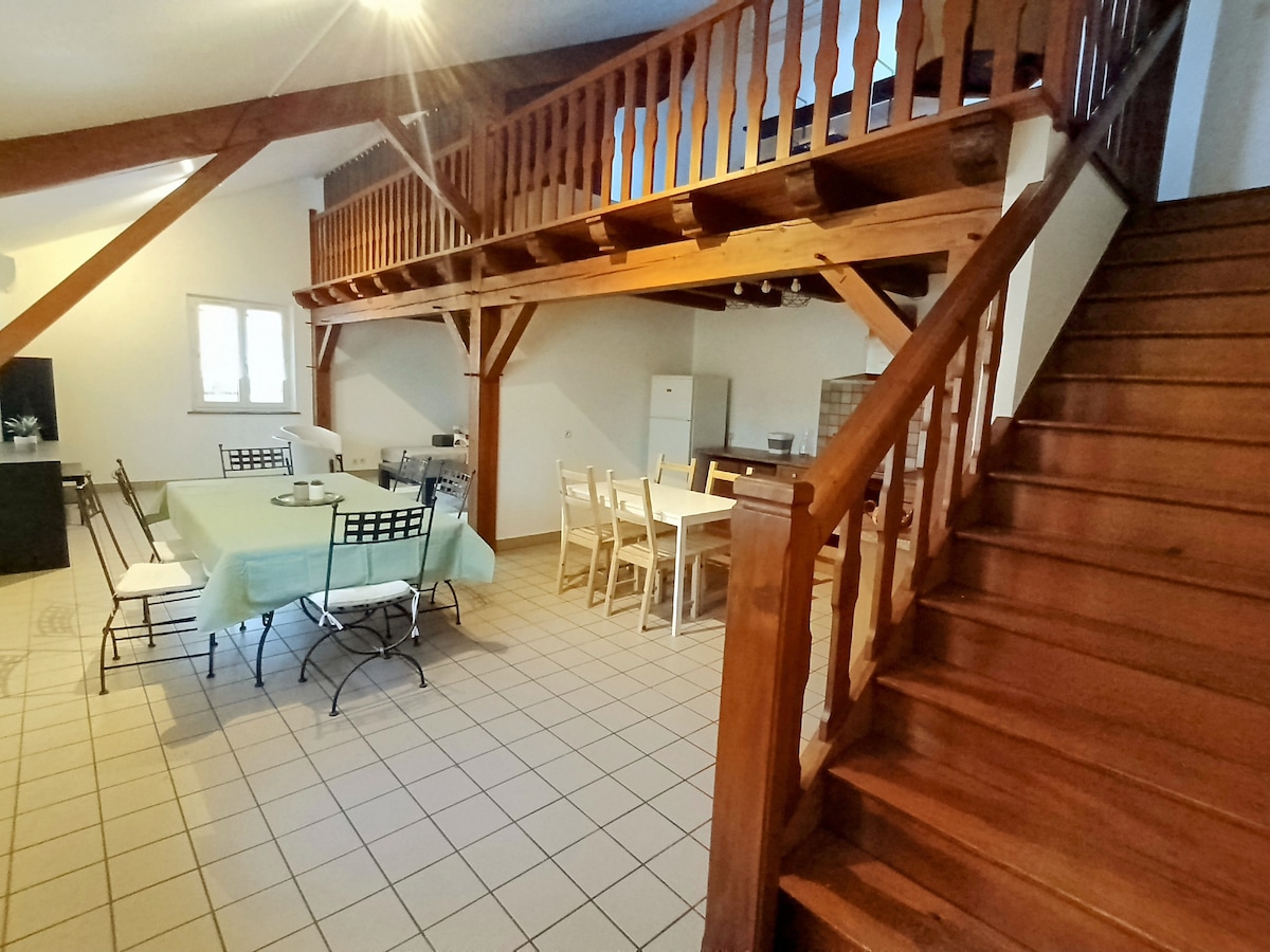 An open living space features wooden beams and a staircase leading to a mezzanine. A large table surrounded by chairs occupies one side, while a smaller dining area is visible. Light streams through windows, illuminating the tiled floor and inviting atmosphere.
