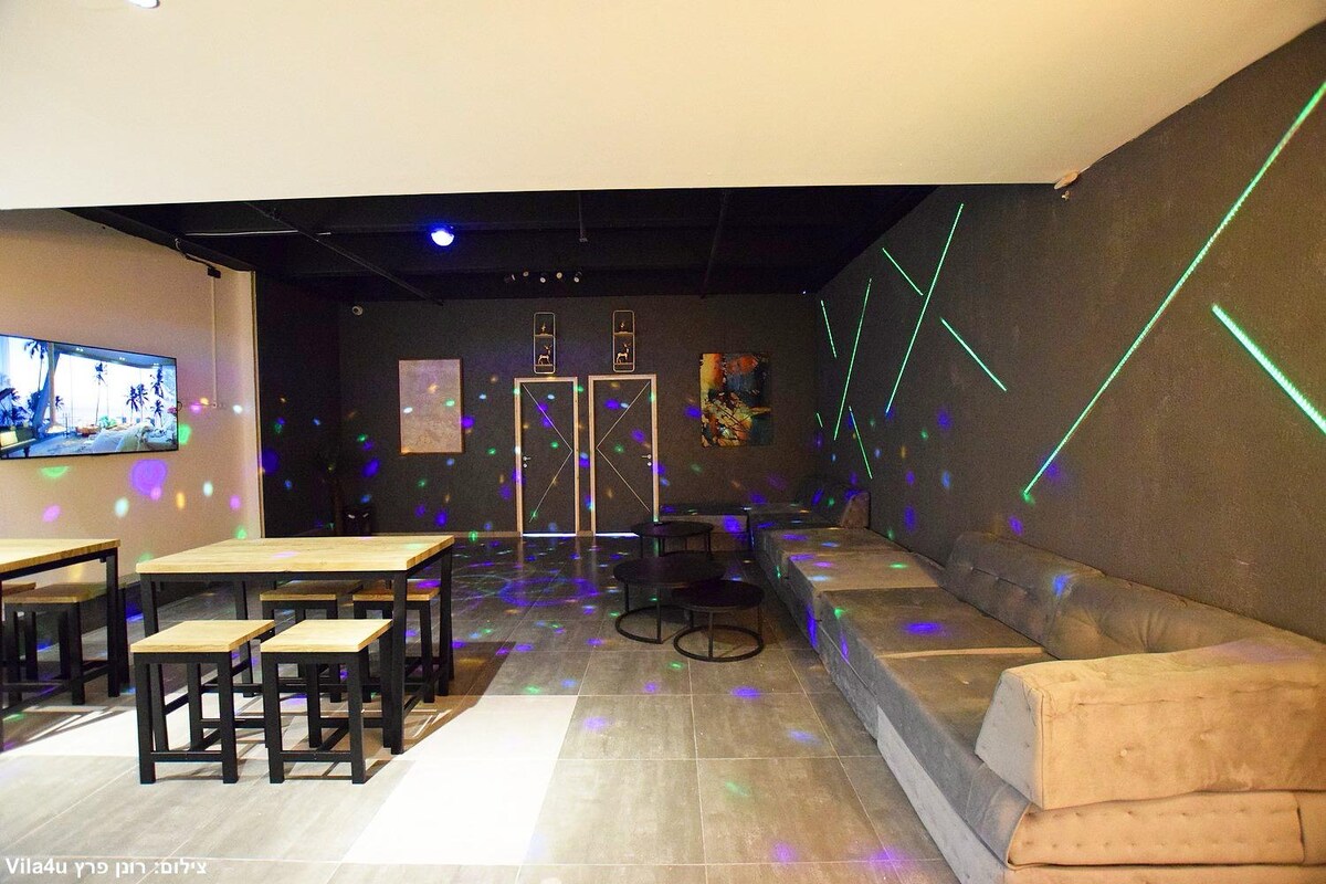 A spacious lounge area is equipped with a large sectional sofa and several seating arrangements. Colorful lights create a dynamic atmosphere on the walls. A large screen is visible on one side, enhancing entertainment options. Natural light from overhead fixtures brightens the scene.