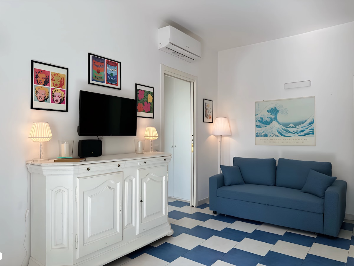 A bright living area features light blue and white checkered flooring. A soft blue sofa is situated against the wall, accompanied by art pieces framed and displayed above. A flat-screen TV rests on a white cabinet, with two bedside lamps providing gentle illumination.