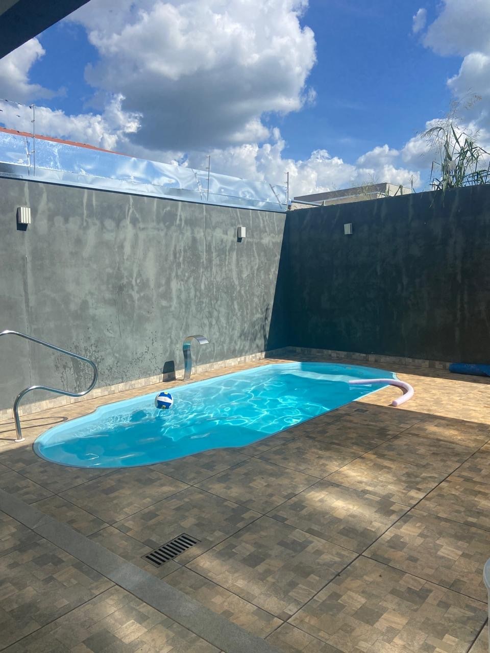 A small swimming pool is situated in a private outdoor area, surrounded by walls that enhance privacy. The water glistens under the bright sky, and a tiled patio provides space for relaxation. Cloud patterns are visible above, adding to the serene setting.