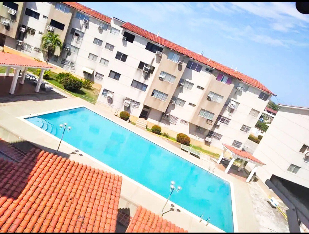 An outdoor swimming pool is centrally located, surrounded by manicured lawns and neatly trimmed bushes. Multi-story buildings are visible in the background, featuring a blend of balconies with shades of beige and grey. A clear blue sky with patches of clouds complements the scene.