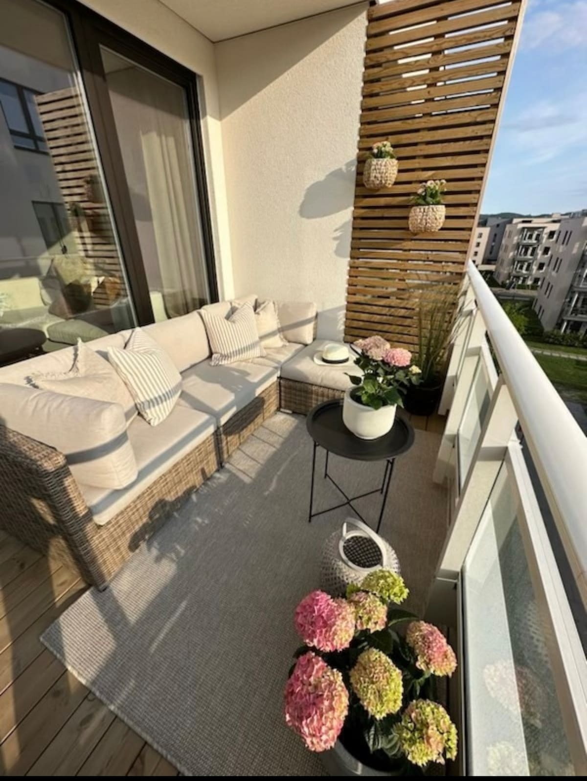 A balcony area features a comfortable seating arrangement with plush cushions arranged around a small table. Decorative plants are placed in pots along with colorful flowers on the floor, enhancing the inviting outdoor space. Natural light filters in, contributing to a serene atmosphere.