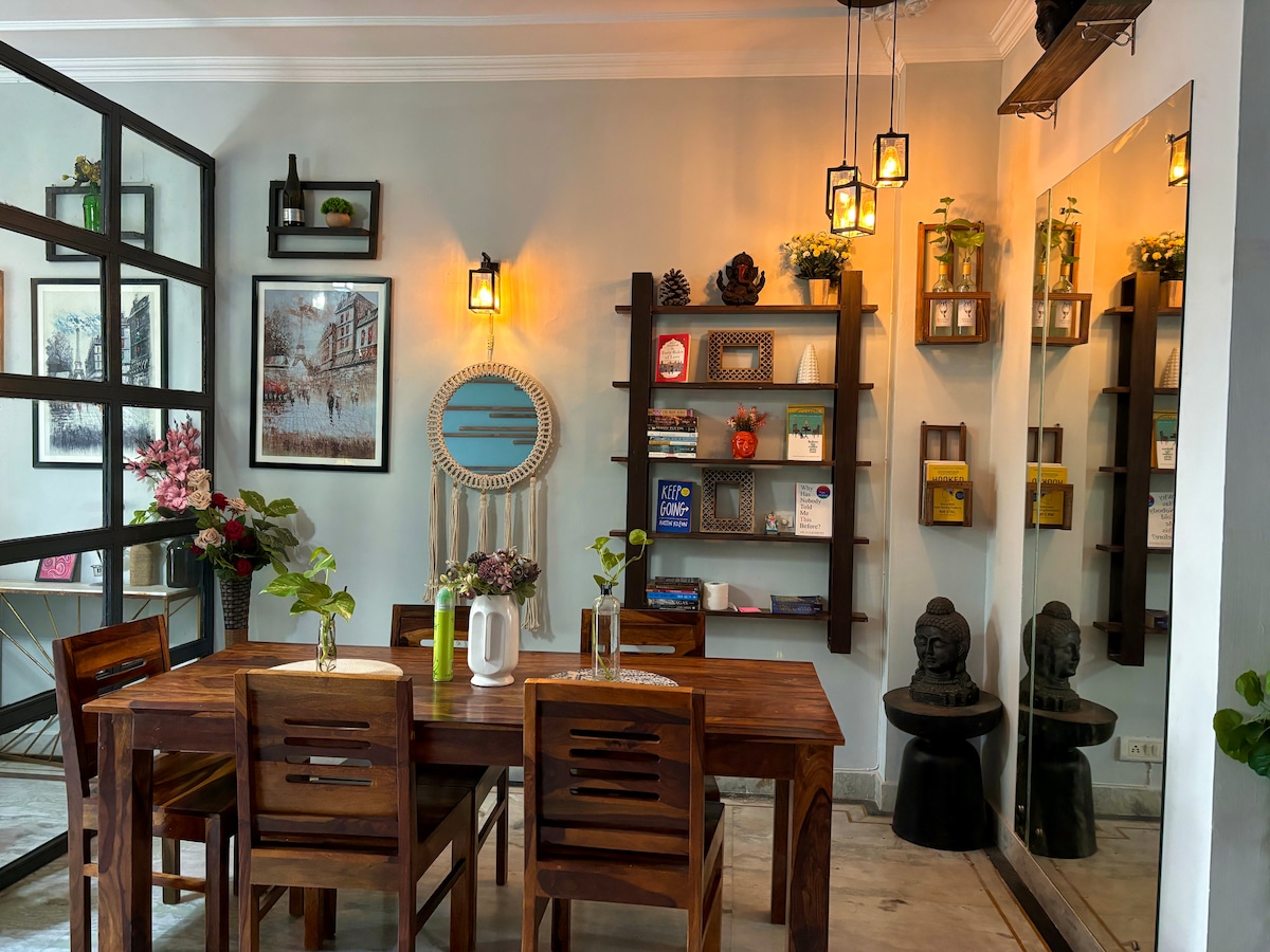 A cozy dining area features a wooden table surrounded by matching chairs. The space is decorated with framed artwork, shelves displaying various items, and a round mirror. Soft lighting from hanging fixtures enhances the inviting ambiance, while plants add a touch of greenery.