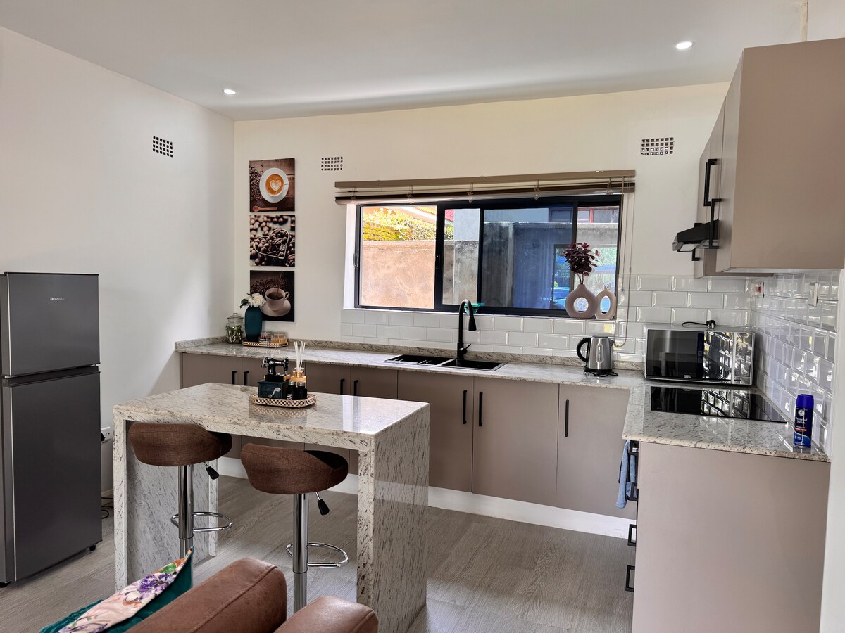 A modern kitchen features sleek cabinetry and a light granite countertop. A window provides natural light, while a small table with two stools sits in the center. Appliances including a refrigerator, kettle, and microwave are neatly arranged, contributing to the functional layout.