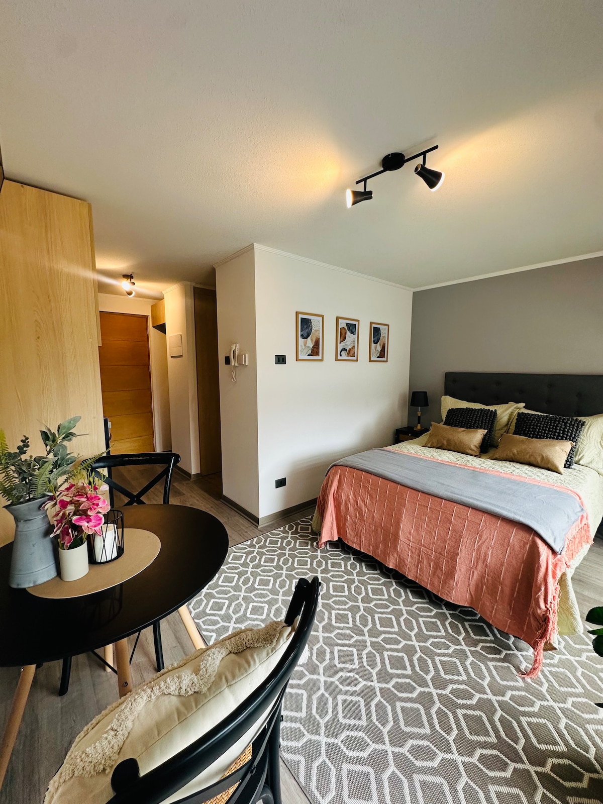 A cozy bedroom is presented with a spacious bed adorned in neutral and warm tones, complemented by decorative pillows. A small dining table and chair provide an area for meals. Artwork is displayed on the wall, and light fixtures enhance the ambiance.