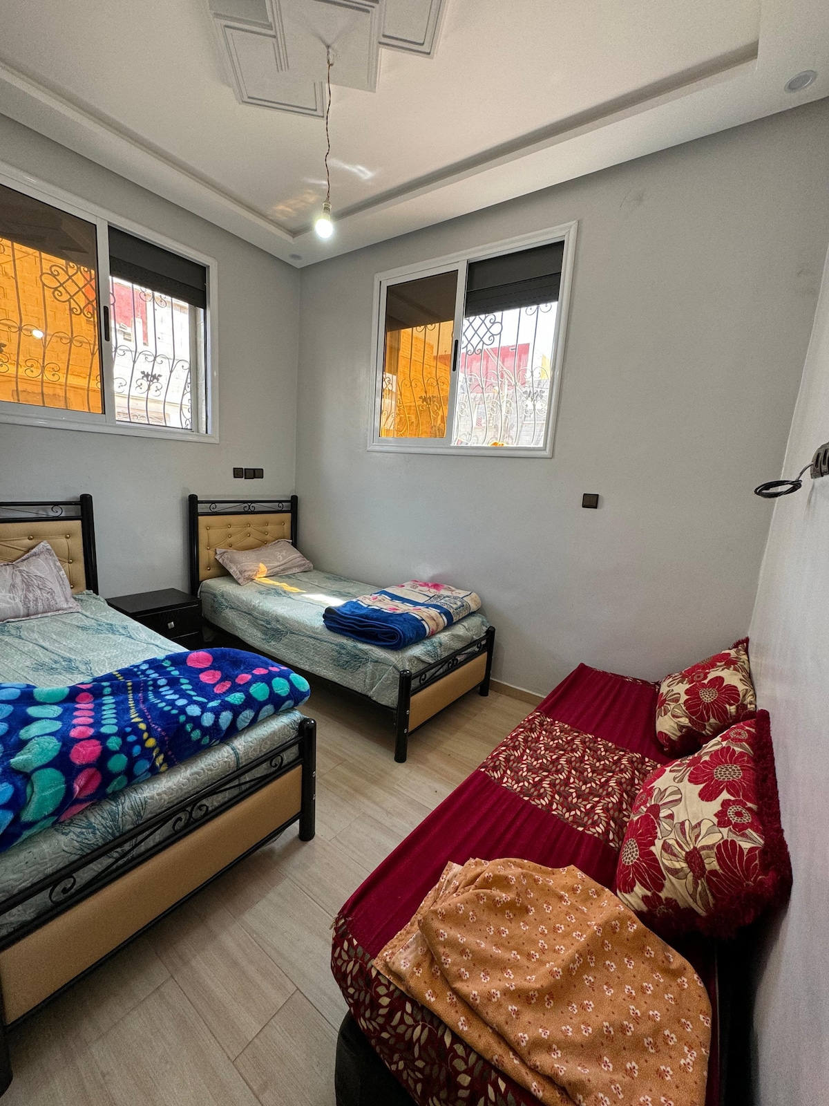 A well-lit bedroom displays two single beds, each dressed in colorful blankets. A third bed, adorned with decorative cushions, is positioned against the wall. Natural light enters through two windows, framed with dark blinds.