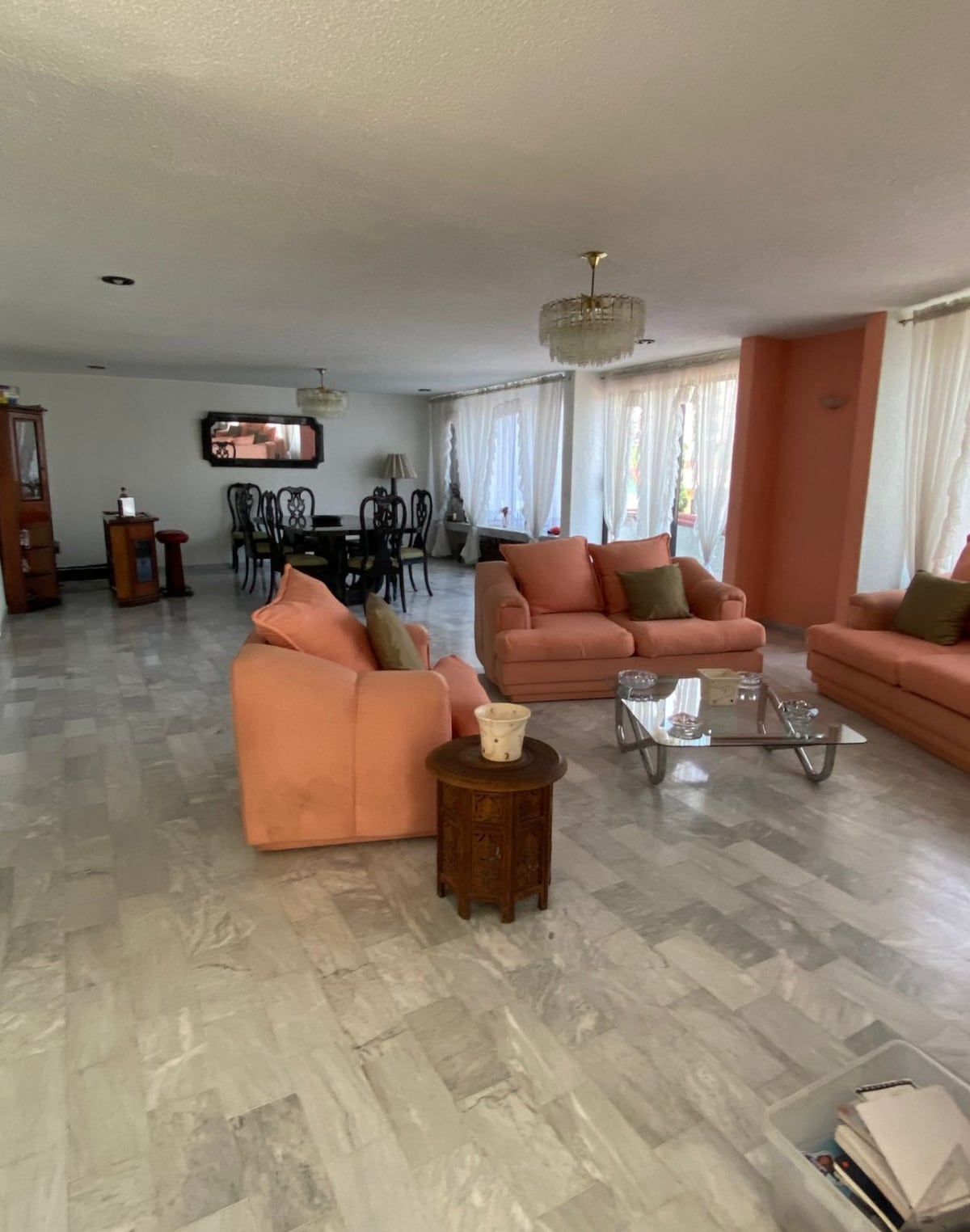 An open living area features light-colored marble flooring and ample natural light from large windows. Two peach-colored sofas, accompanied by decorative cushions, are arranged around a glass coffee table. A dining area with black chairs and a dark wooden table is visible in the background.