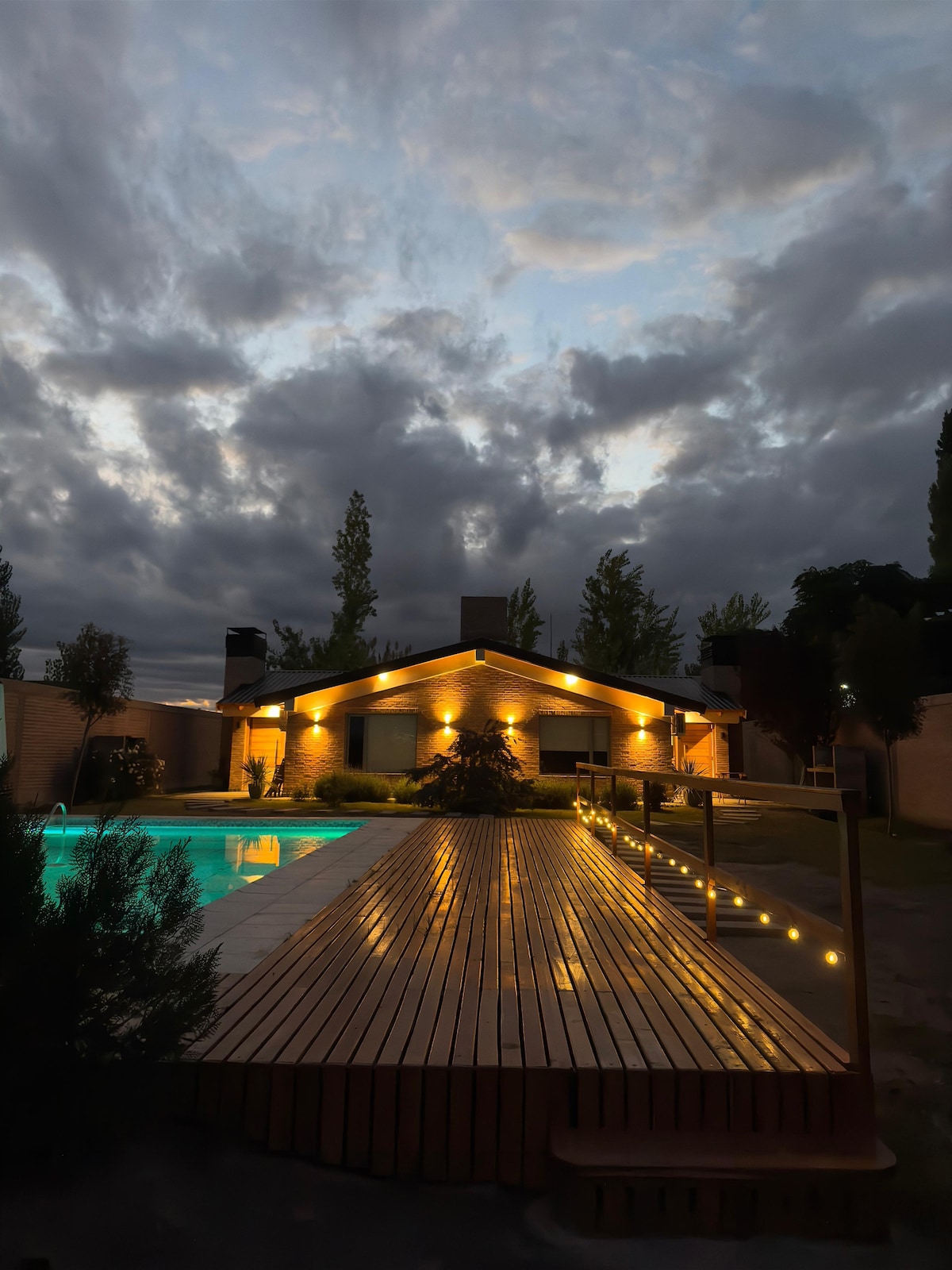 A tranquil outdoor space features a lit wooden deck leading to a sparkling pool. The residence showcases a warm ambiance with soft lighting illuminating the exterior walls, and lush greenery complements the inviting environment as clouds drift across the evening sky.