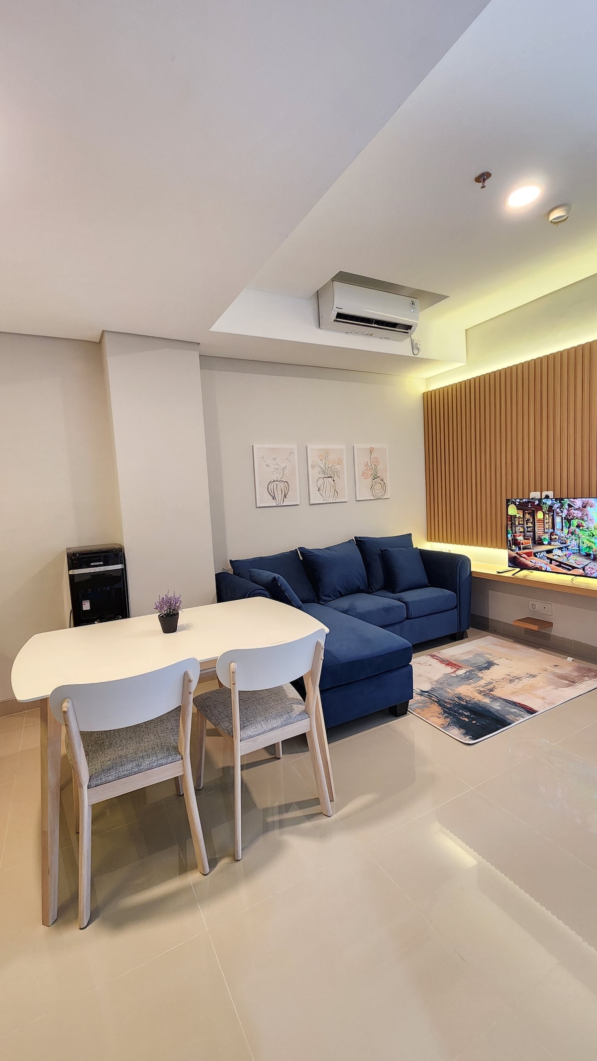A comfortable living space features a blue sofa facing a mounted television. A round dining table with two chairs is placed nearby. Light-colored walls are complemented by a wooden accent wall, and framed artwork adds a decorative touch to the area.