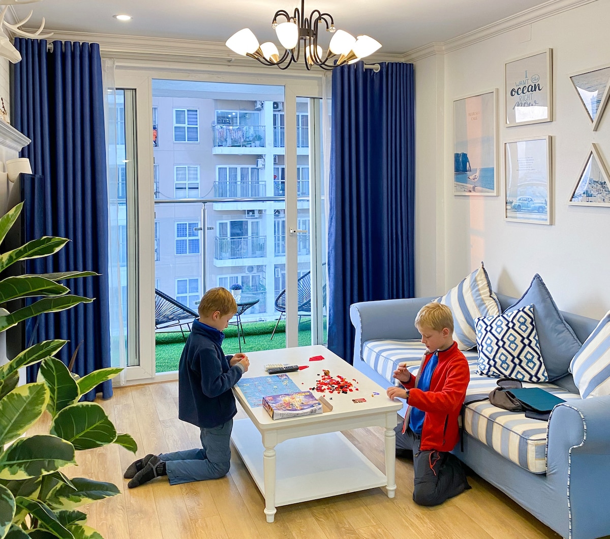 A bright living area is presented with a comfortable blue couch adorned with decorative pillows. A small white table is set in the center, with two children engaged in a game. Large glass doors provide views of the outside balcony, framed by navy curtains.