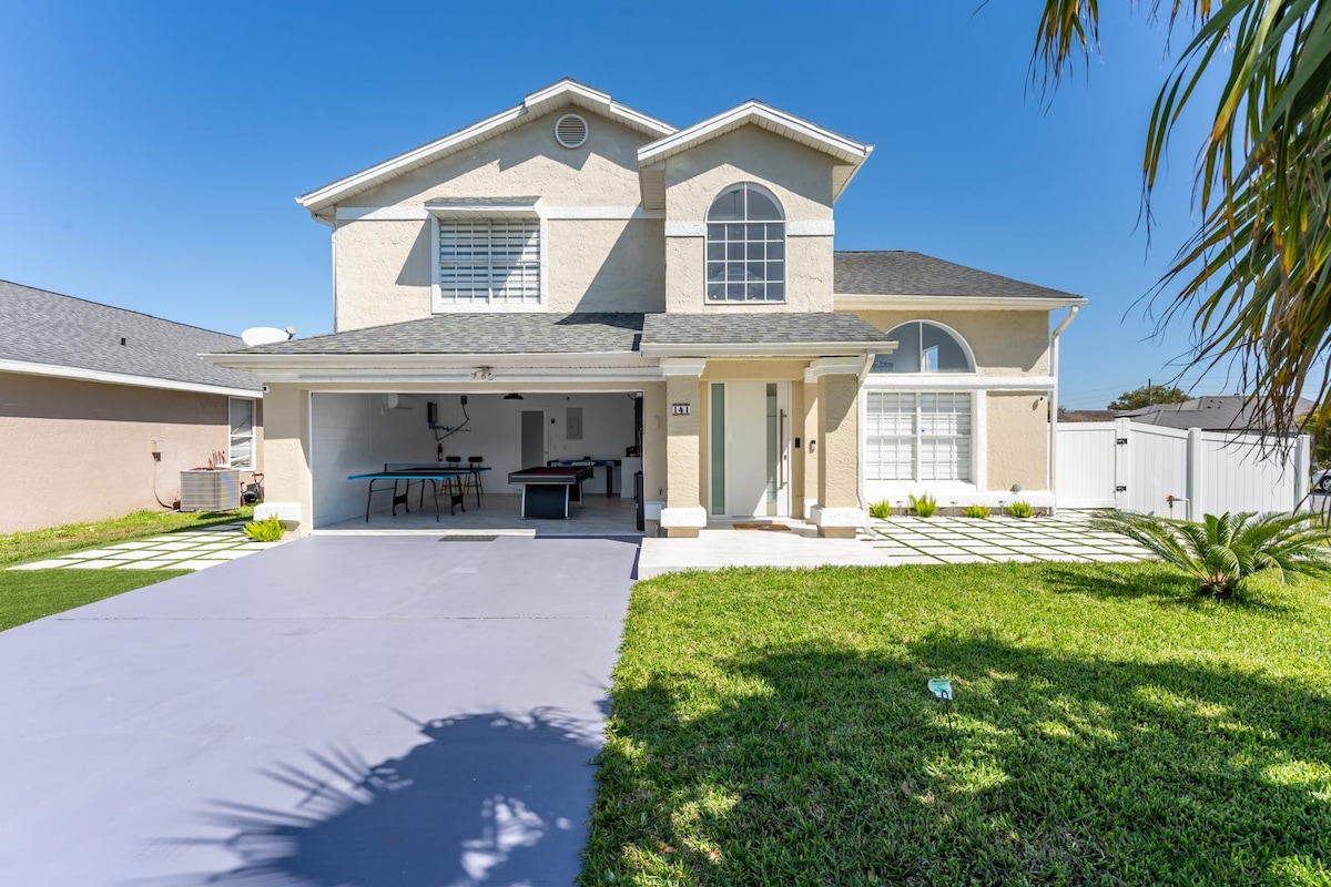A modern two-story house is showcased, featuring a bright exterior and a spacious driveway. Lush green grass surrounds the property, while palm trees add a tropical touch. Large windows on the upper level allow for ample natural light, highlighting the clean lines of the architecture.