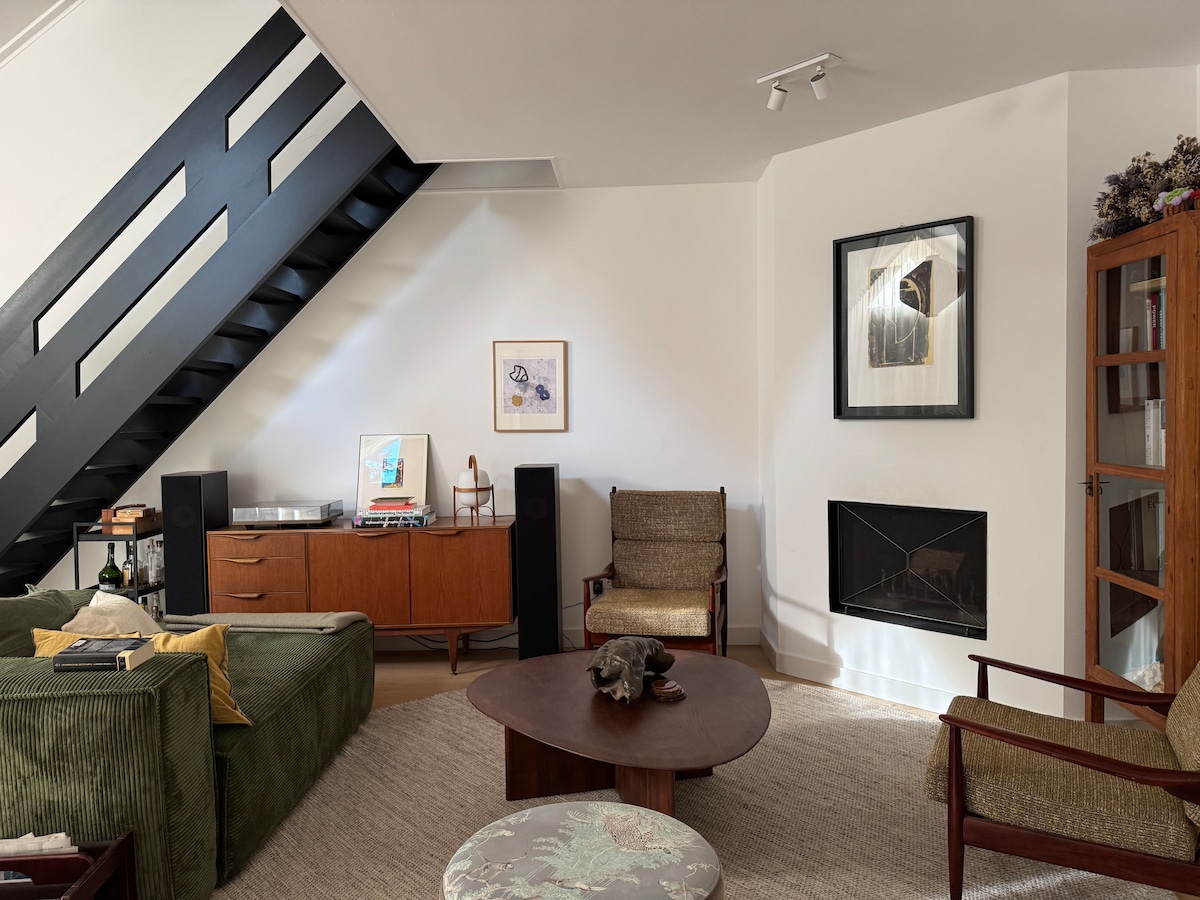 A living area features a green upholstered sofa and two armchairs arranged around a round coffee table. A modern sideboard displays audio equipment, while a fireplace is positioned between artistic wall frames. The room is enhanced by sloped ceilings and natural light from nearby windows.