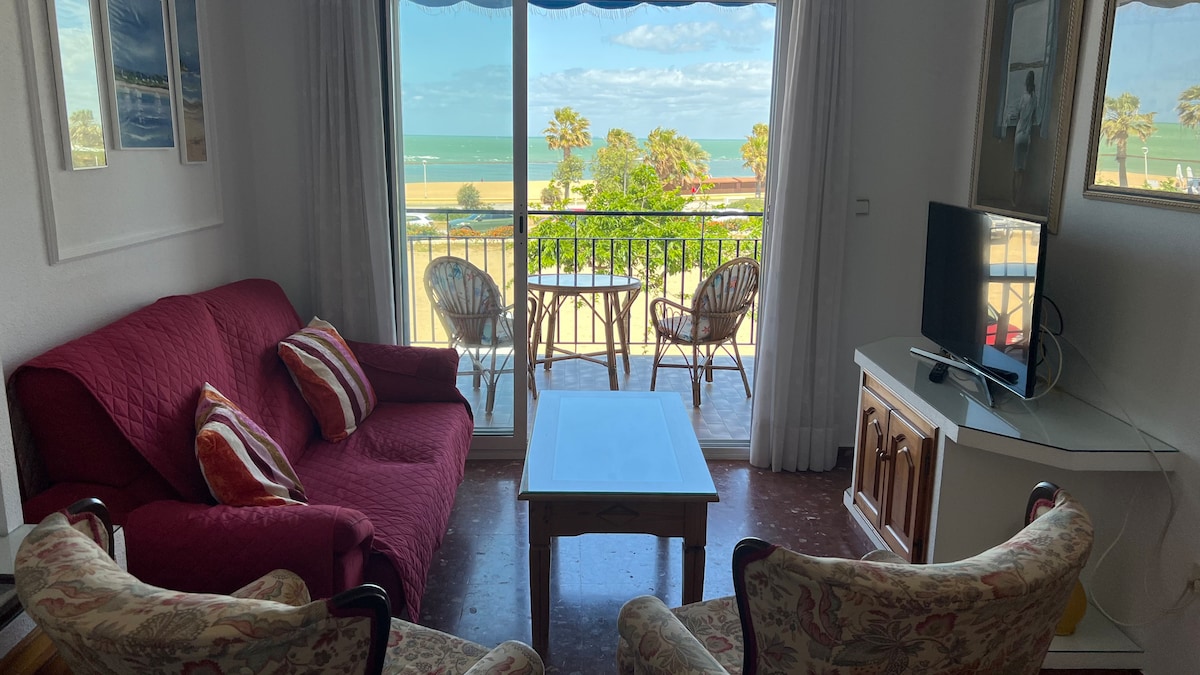 A living area features a maroon sofa with striped cushions and two upholstered armchairs. A white coffee table sits in front, with a view of the balcony showcasing two chairs and a table. The sea is visible in the background through large glass doors, allowing natural light.