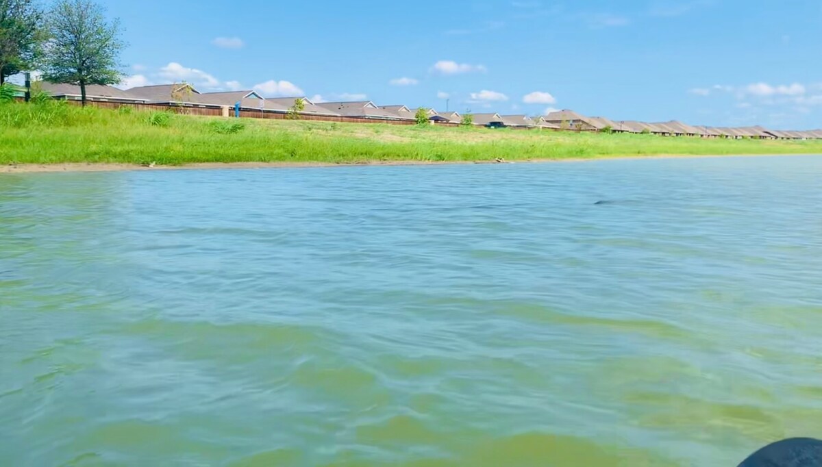 A calm man-made lake is showcased with clear, gentle waters reflecting a blue sky. In the background, a series of houses line the shore, and green grass frames the lake's edge, adding a serene and natural touch to the setting.
