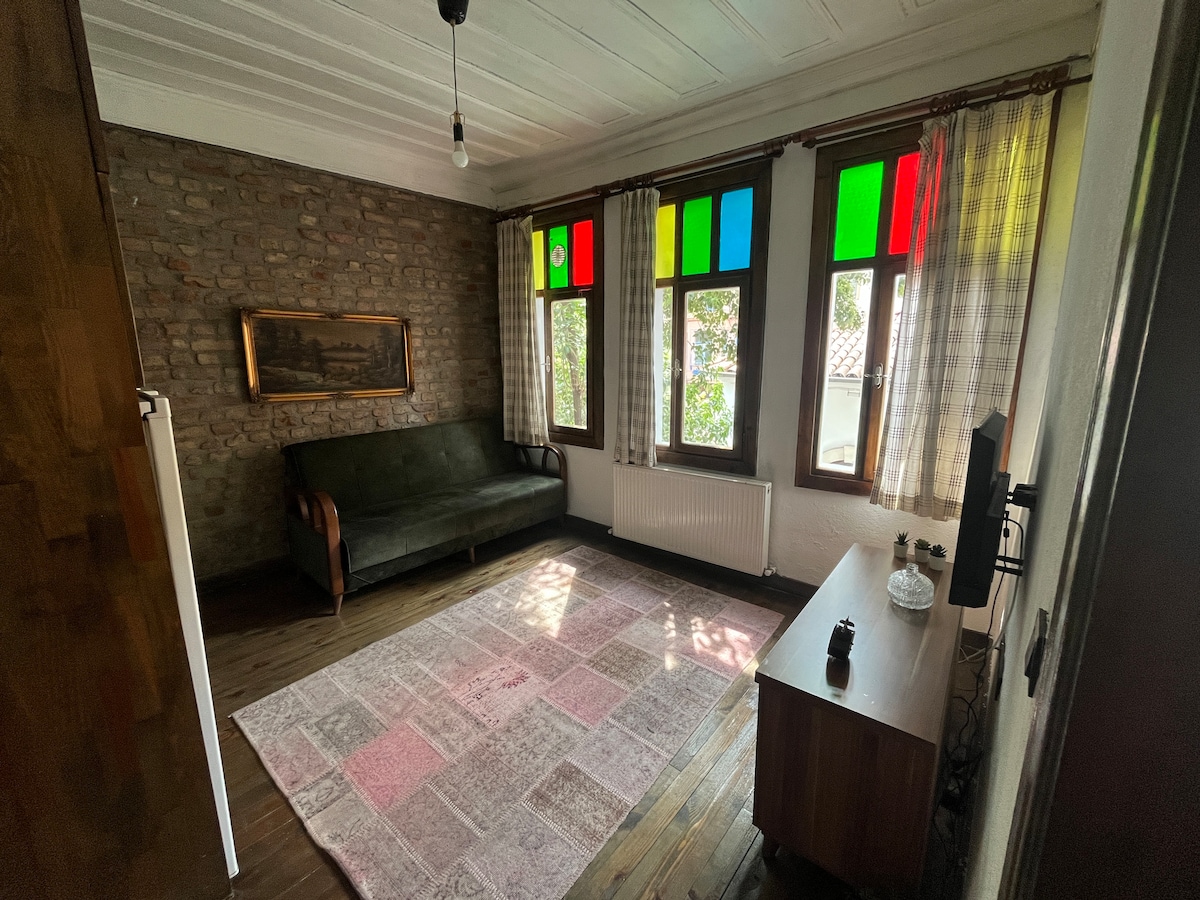 A living area features a dark green sofa positioned against a brick wall, complemented by colorful stained glass windows allowing natural light to filter in. A light-colored area rug covers the wood floor, while a wooden cabinet is placed against one wall.