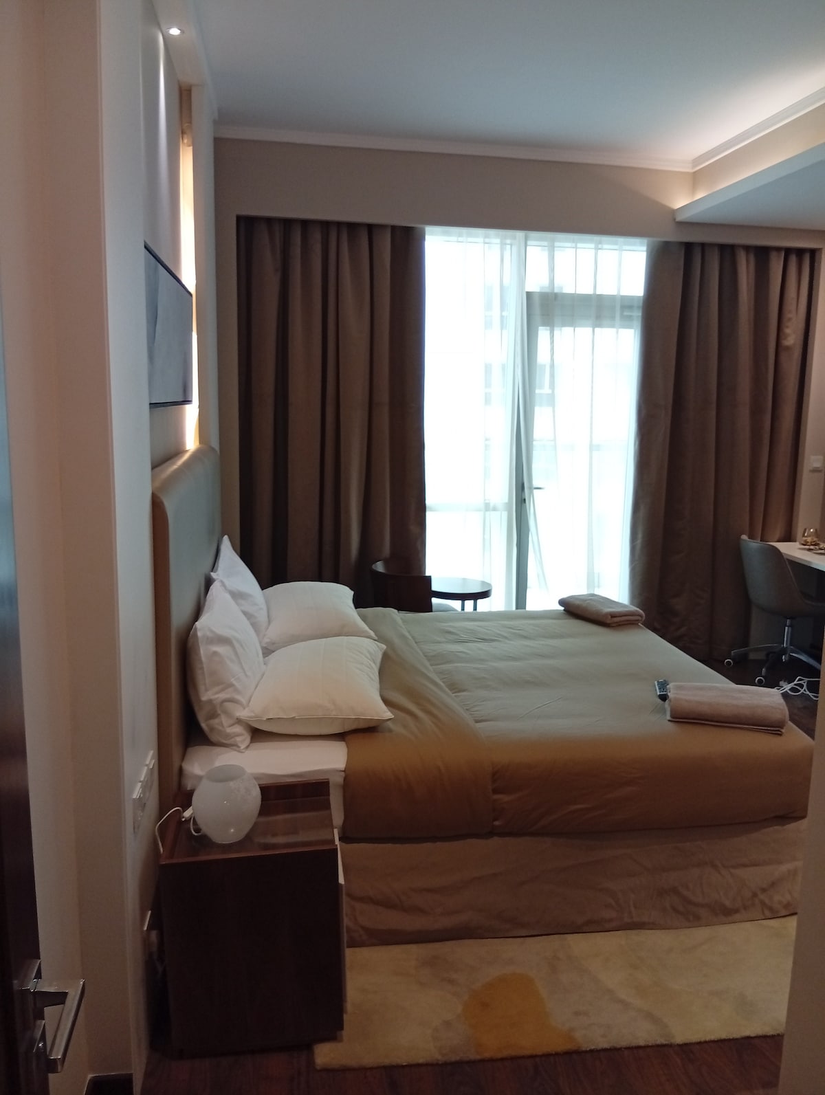 A cozy bedroom features a spacious bed with neutral-toned linens and two folded towels on its surface. Natural light filters in through sheer curtains, illuminating the light wood furnishings and a small side table. A desk is positioned nearby against the wall.