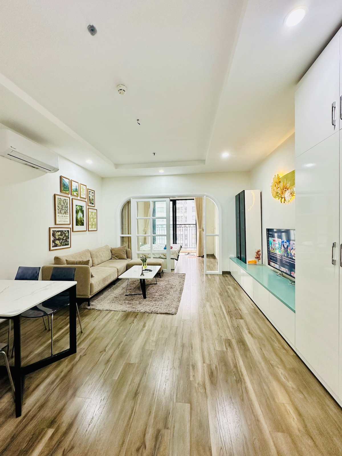 A modern living area is presented with a comfortable sofa and a spacious coffee table, surrounded by framed artwork on the walls. A large window offers natural light, leading to a balcony. A flat-screen TV is mounted on the sleek cabinet, enhancing the overall functionality.