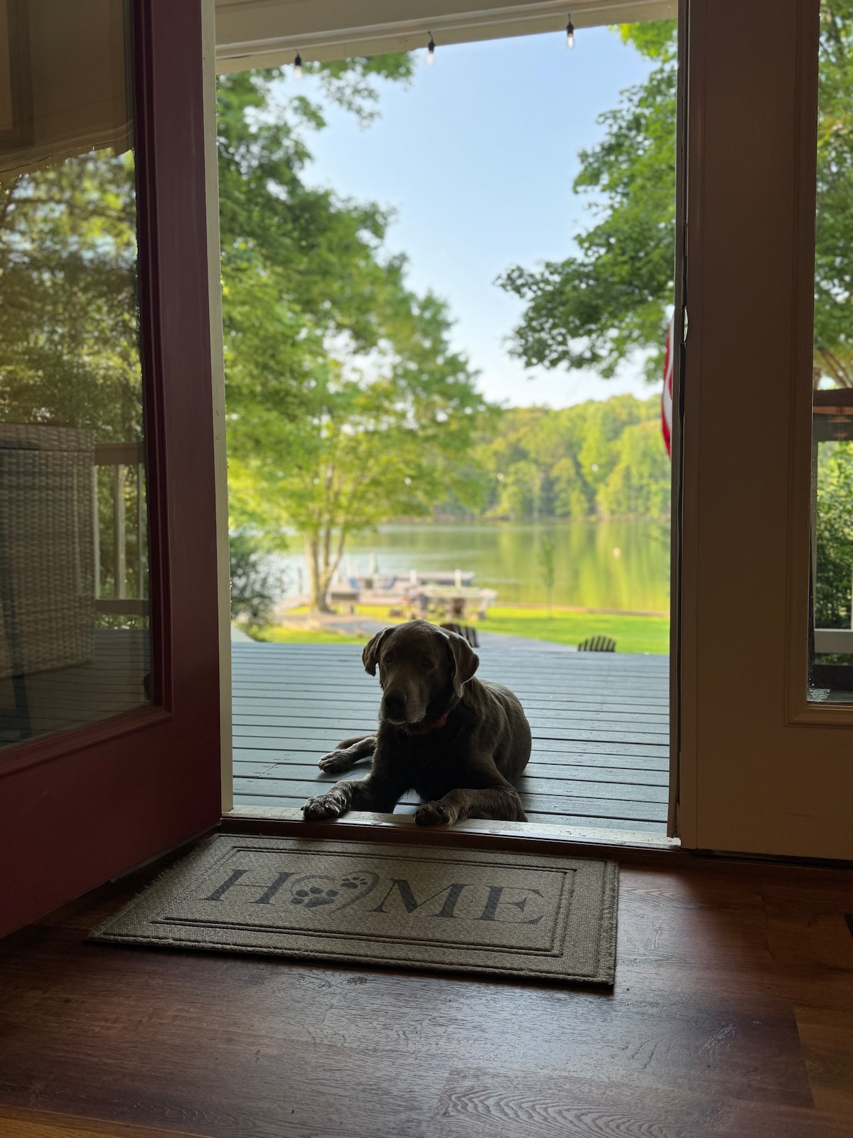 A welcoming entrance features a cozy doormat with the word 'HOME'. A dog rests on the porch, with views of the serene lake and surrounding greenery visible in the background.
