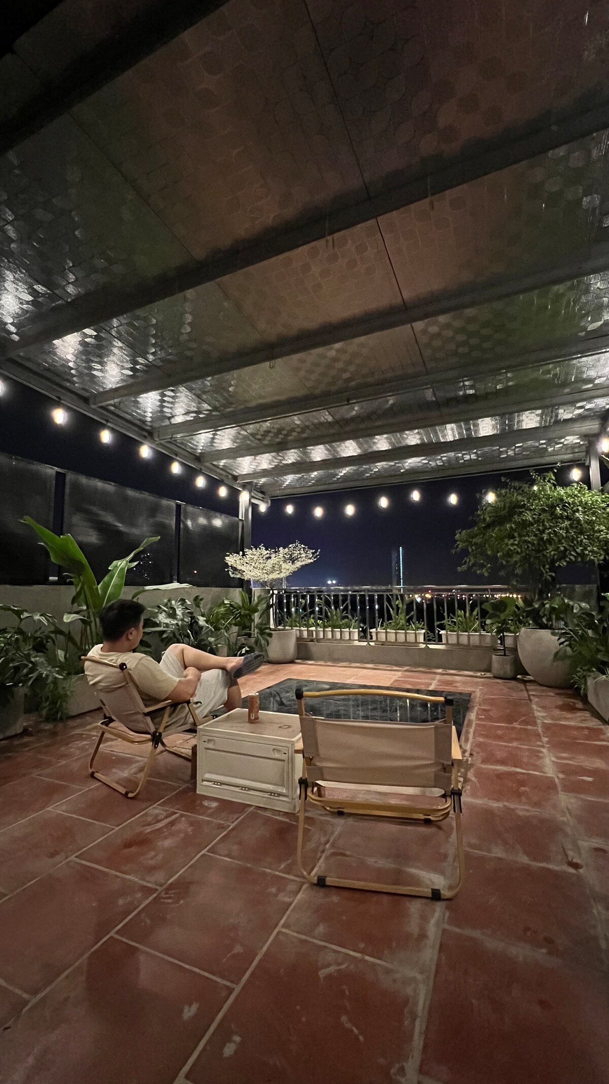 An outdoor rooftop area is featured with a small pool surrounded by potted plants. Two lounge chairs are positioned on a tiled surface, and string lights provide soft illumination in the evening. The view is open, connecting to the surrounding night sky.