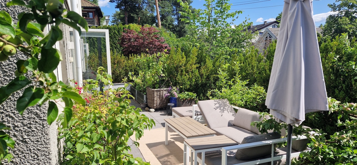 An outdoor terrace is depicted, featuring a lounging sofa and a table. A white umbrella provides shade, surrounded by lush greenery and vibrant plants. The area offers a peaceful spot for relaxation, with a paved surface and a tranquil ambiance created by the surrounding foliage.