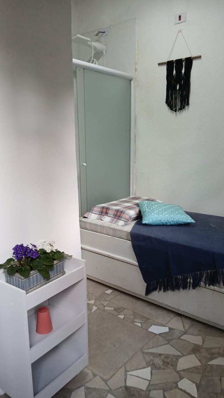 A compact sleeping area is featured, including a stylish bed dressed with varied cushions and a blanket. A small shelf stands nearby, adorned with blooming plants. Natural light filters through frosted glass, highlighting the neutral-colored walls and textured floor.