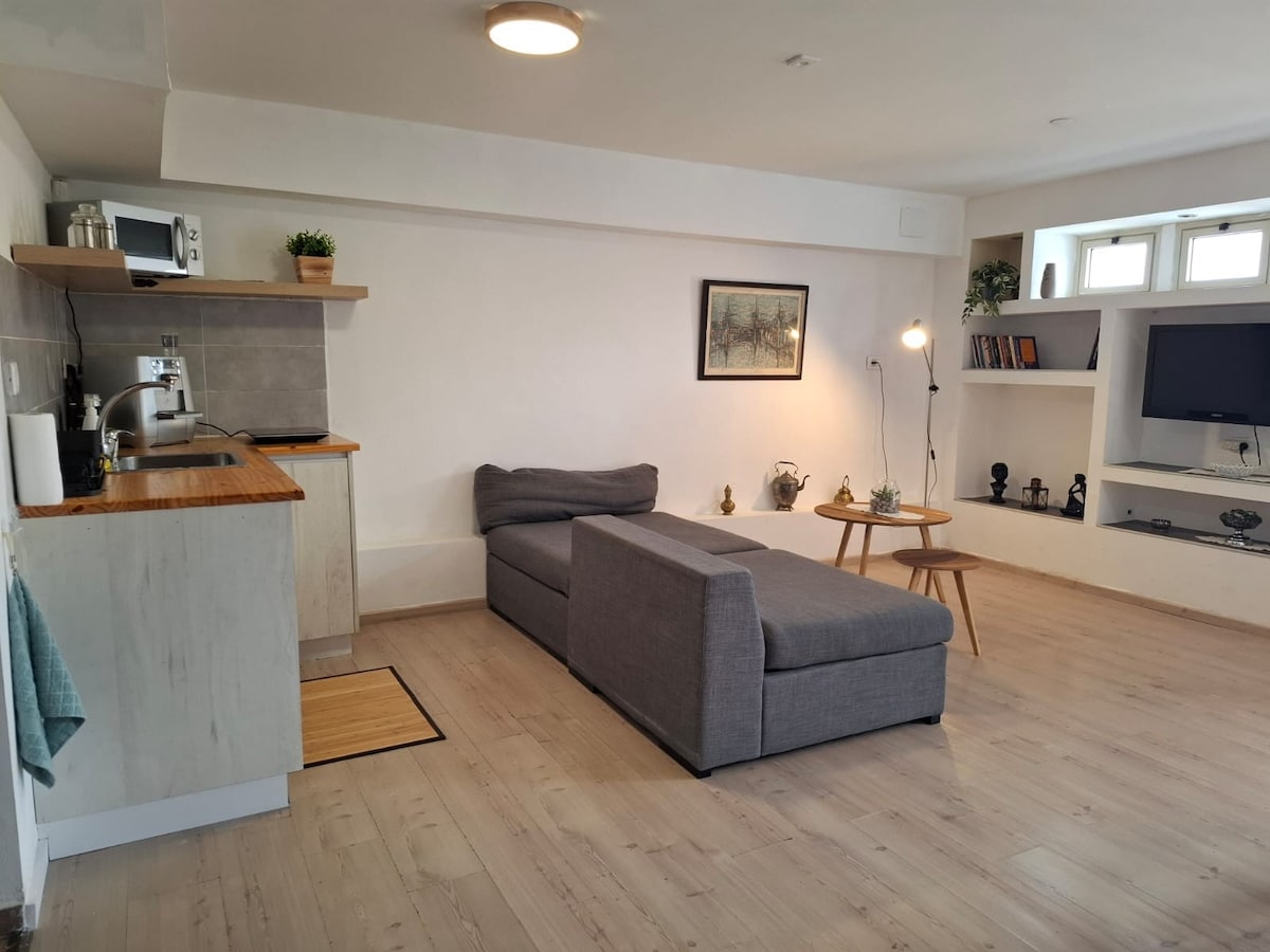 A bright living space is featured, showcasing a cozy gray sectional sofa positioned beside a light wood coffee table. A small kitchenette is observed with essential appliances, while light pours in from windows set within the clean, minimalist design.