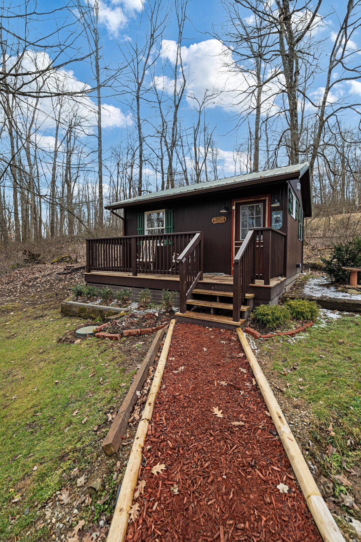 A charming cabin is set amongst tall trees, featuring a simple wooden deck with steps leading to a welcoming entrance. A red mulch pathway guides visitors from the grassy yard to the front door, enhancing the connection to nature.