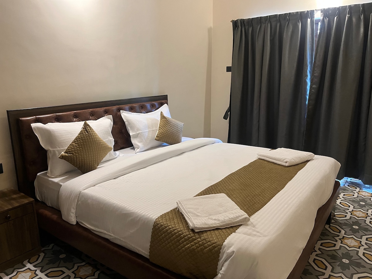 A well-appointed sleeping area features a spacious bed with elegant bedding and decorative cushions. Two towels are neatly arranged at the foot of the bed. Natural light filters through a window, with dark curtains providing privacy. The floor displays intricate tile patterns.