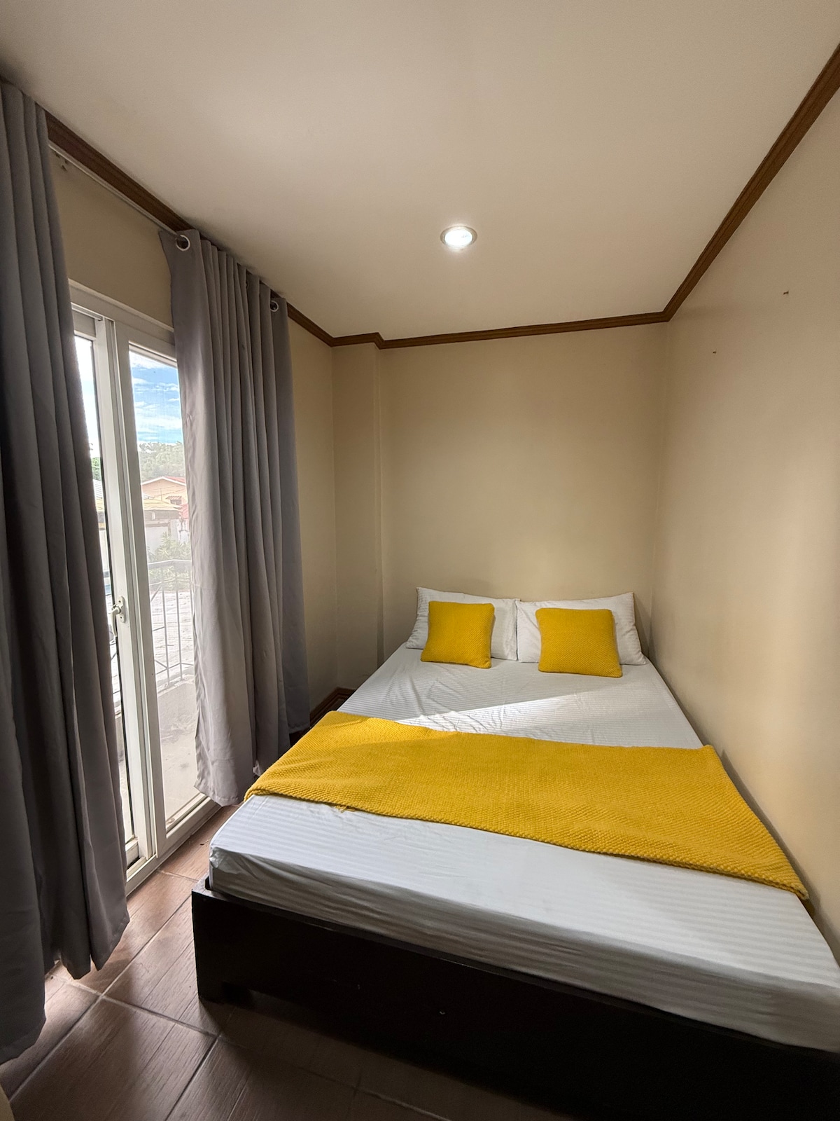A small sleeping area features a spacious bed with white linens and two yellow accent pillows. A bright yellow throw blanket is draped across the bed. Natural light illuminates the room from a nearby balcony door, with light grey curtains framing the window.