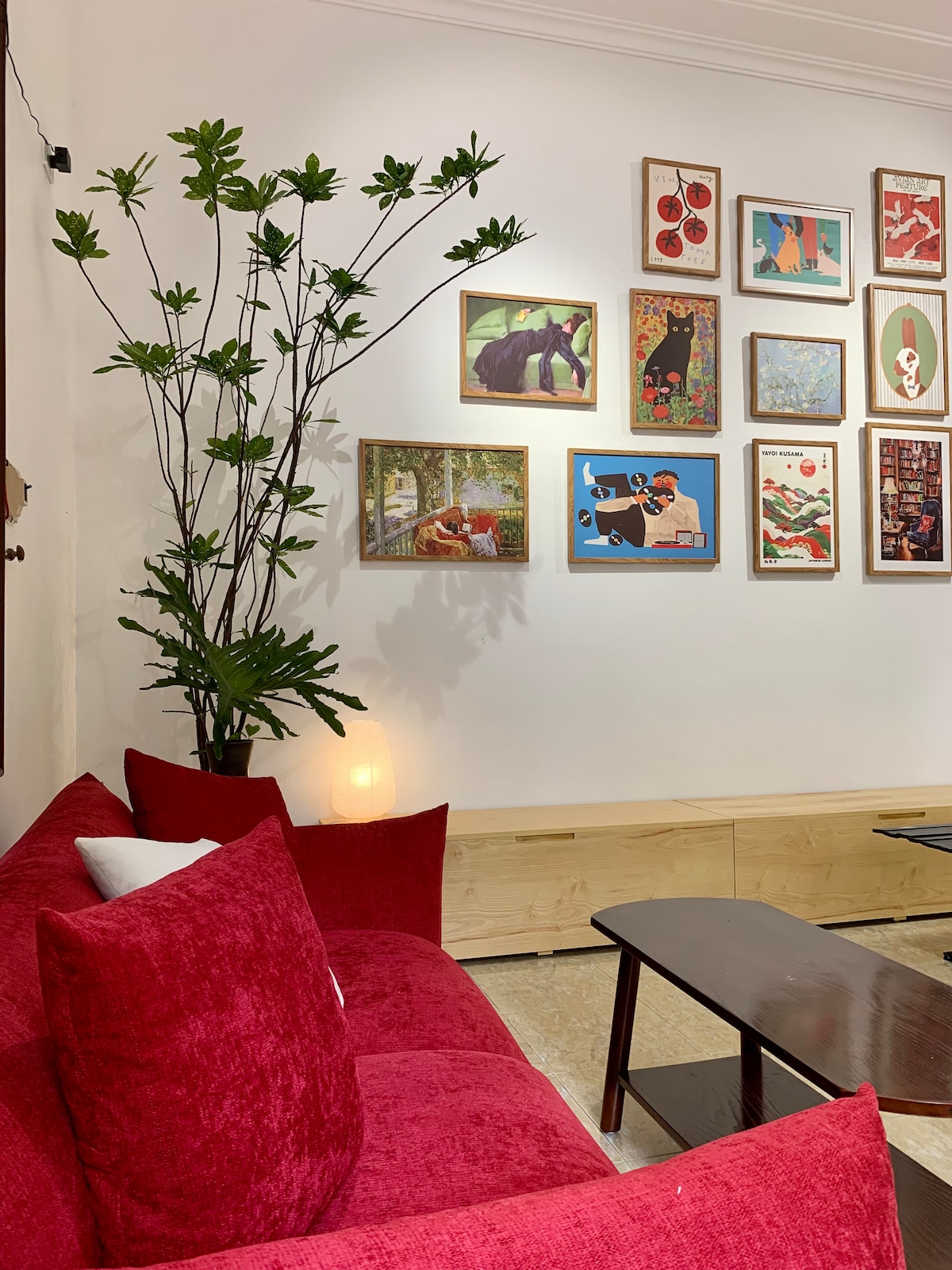 A cozy seating area features a vibrant red sofa adorned with multiple cushions. A dark coffee table sits in front, while a tall indoor plant adds a touch of greenery. An array of framed artwork is displayed on the wall, contributing to the inviting ambiance.