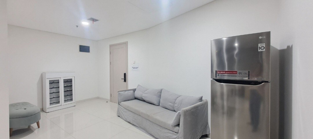 A living area is shown with modern, minimalist design elements. A light gray sofa is positioned against a plain wall, accompanied by a small teal ottoman. A stainless steel refrigerator stands to the right, while a wine cooler occupies the opposite side, enhancing the functional layout.